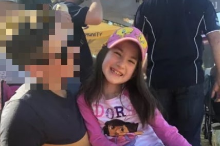 An eight-year-old girl with dark hair and wearing a pink cap sits on the knee of a person whose image is blurred.