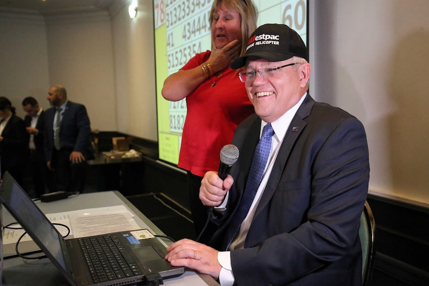 Scott Morrison holds a microphone as he sits at a computer smiling while calling the bingo