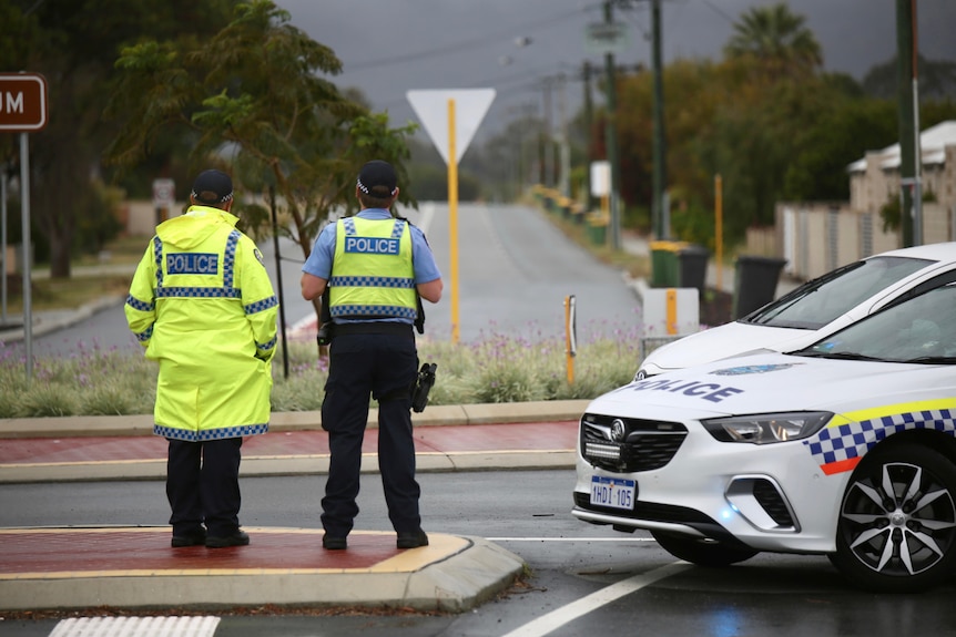 Two police officers stand with their backs turned next to a police car on a road.