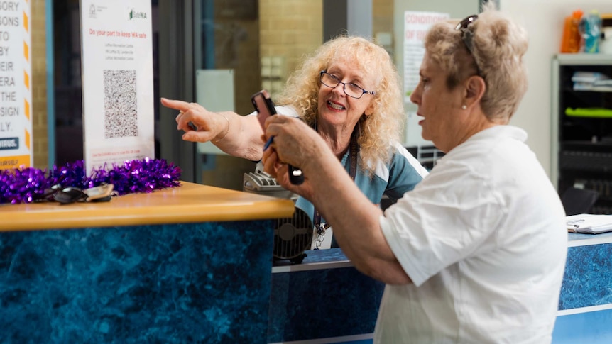 Two women at a counter, one using a phone to scan a QR code while the other points to it.