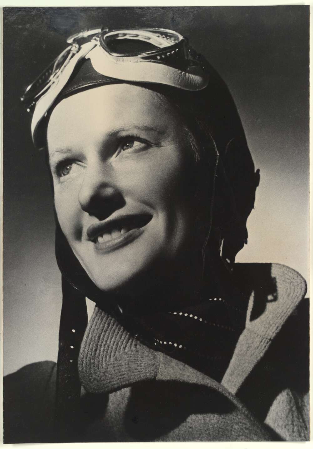 A black and white portrait of a woman in flying hat and goggles smiling looking up to the light.