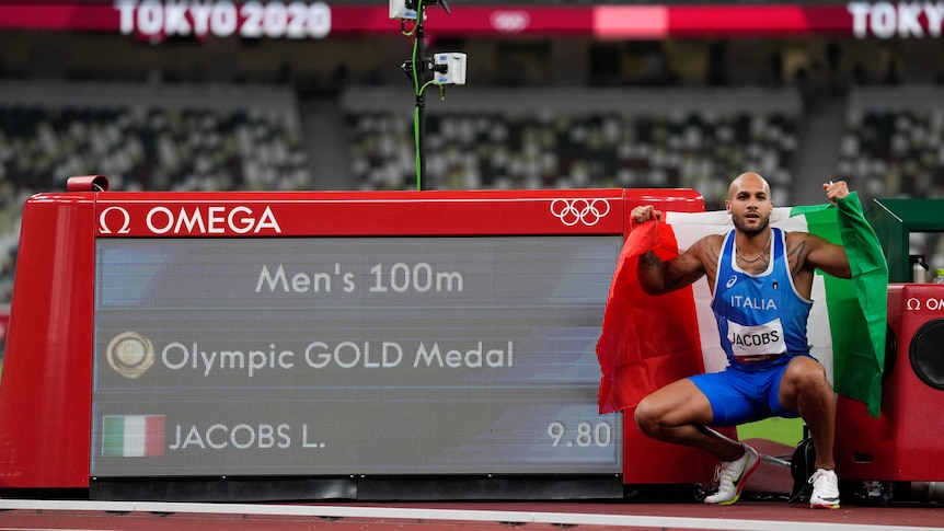 Italian sprinter Lamont Marcell Jacobs squats next to a screen showing his time after the men's 100m final at Tokyo Olympics.