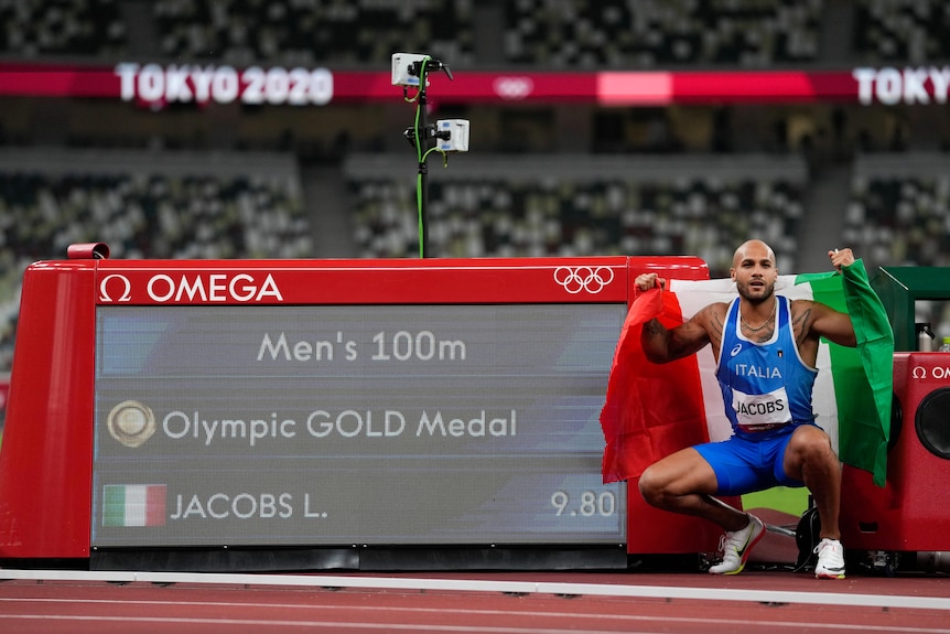 Italian sprinter Lamont Marcell Jacobs squats next to a screen showing his time after the men's 100m final at Tokyo Olympics.