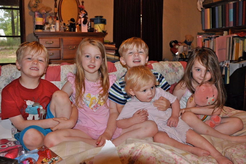 Five kids with light blonde hair sit together on a couch in pyjamas.