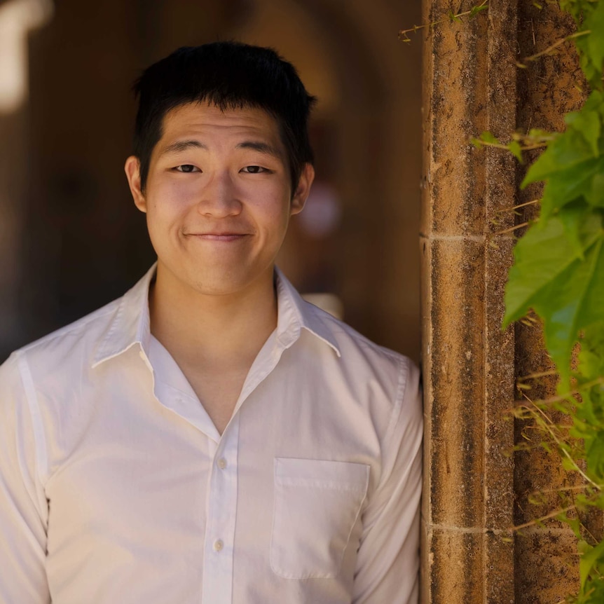 A young Asian man smiling at the camera wearing a white shirt with some vines growing to the right of him
