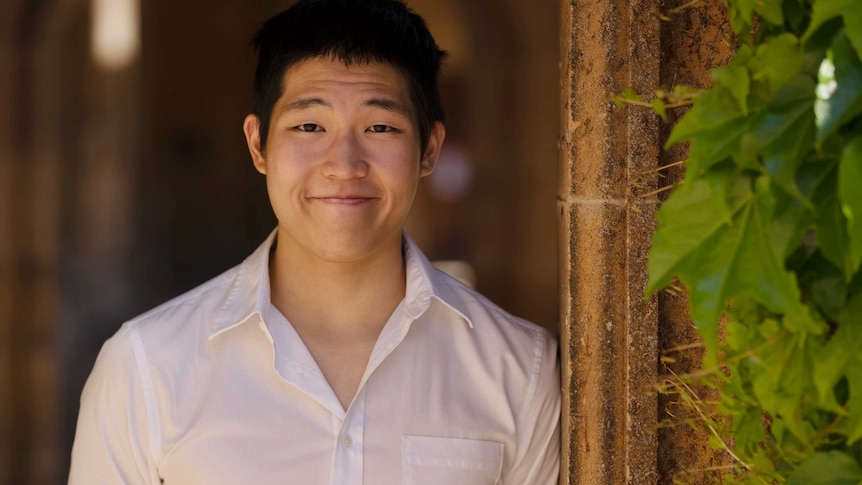A young Asian man smiling at the camera wearing a white shirt with some vines growing to the right of him