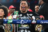 Devin Haney smiles while holding a number of title belts inside a boxing ring