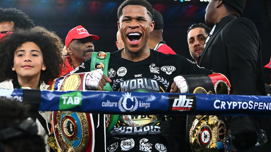 Devin Haney smiles while holding a number of title belts inside a boxing ring