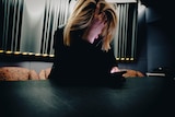 A woman dressed in black with blond hair looks at her phone, fave in her hand