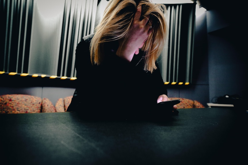 A woman dressed in black with blond hair looks at her phone, fave in her hand
