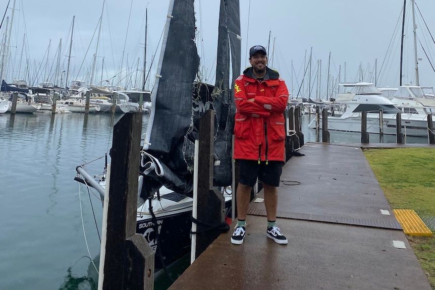 A man stands in gloomy weather in front of a boat with a ripped sail