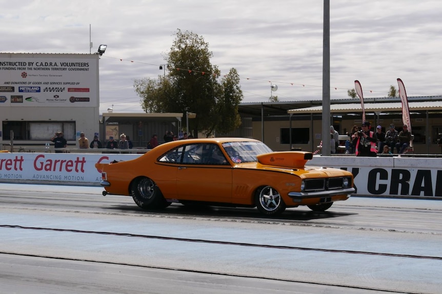A photo of a unique, orange car on a race track. Crowds are looking.