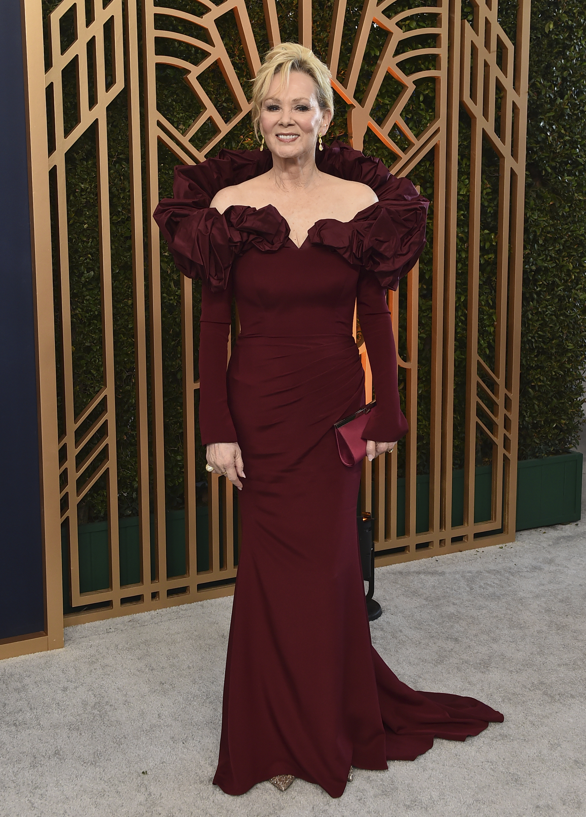 Jean smart wears a maroon floor length gown with long sleeves and a lot of ruffling around the shoulders. 