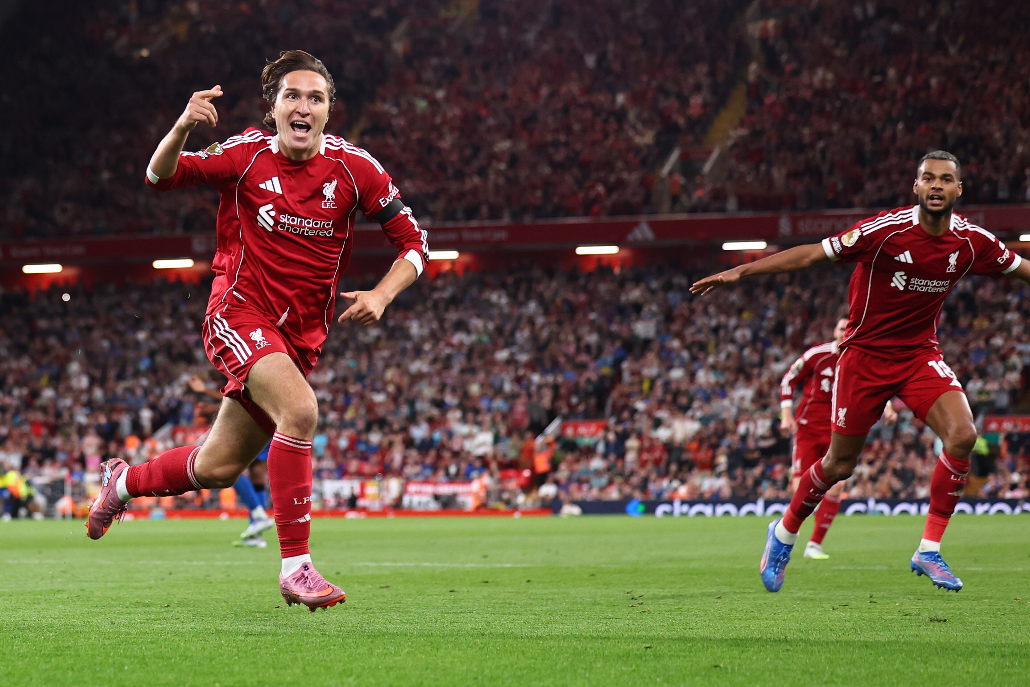 A Liverpool player points and yells in celebration as he runs away from goal, and a teammate looks on in the background.