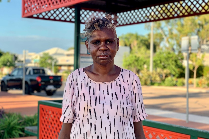 A dark-skinned woman stands alone outside, staring directly at the camera.