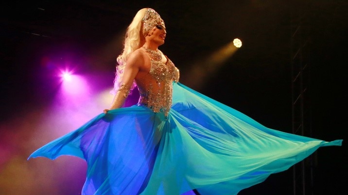 a drag queen singing on stage is a large flowing dress with lights behind her