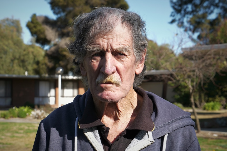 A man with grey hair and a moustache wears a jumper and frowns at the camera. 