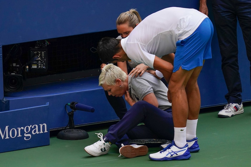 A tennis player bends over to talk to a woman sitting on the ground with her hand to her throat.