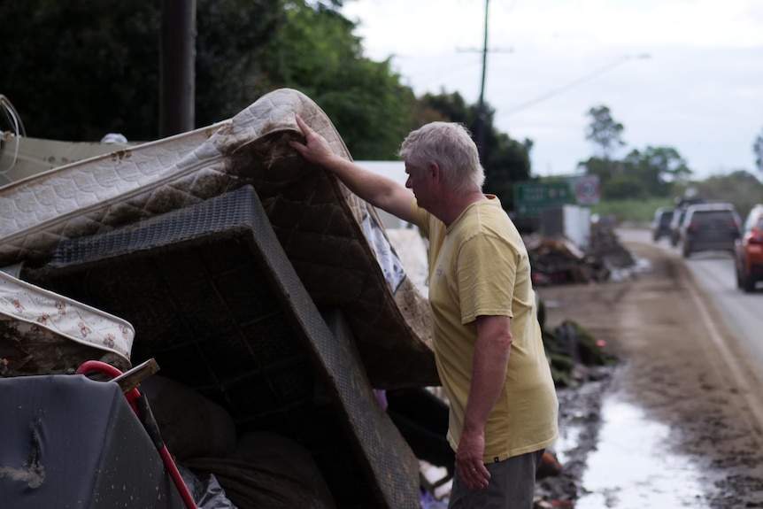 A man in a flooded, muddy street, examines a pile of mattresses. 