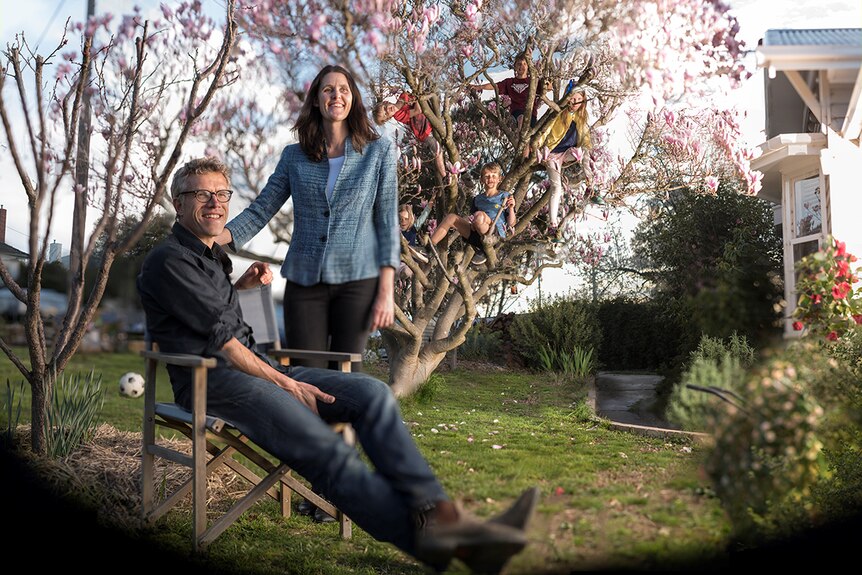 Husband and wife in front of a flowering tree, with five children climbing the tree.