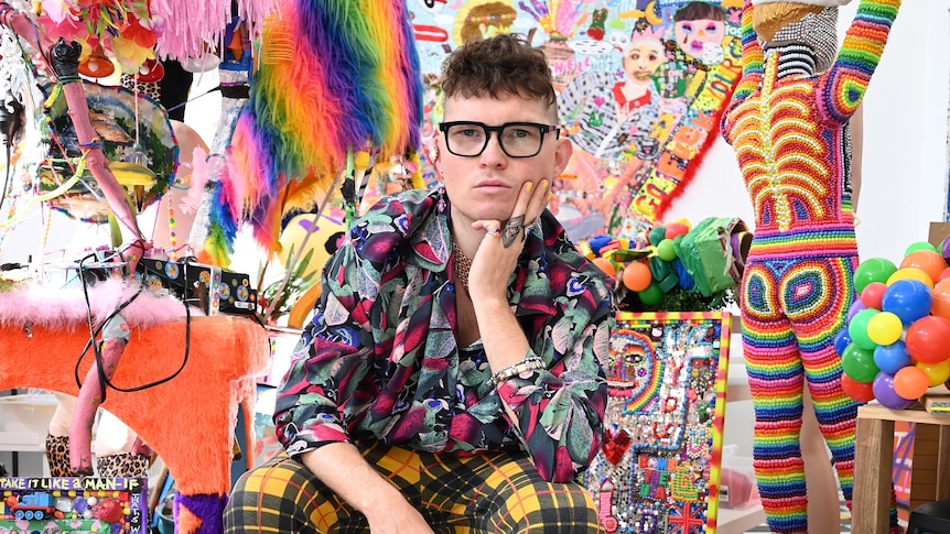 A young man wearing garish clothes and black-framed glasses sits amongst his colourful artwork