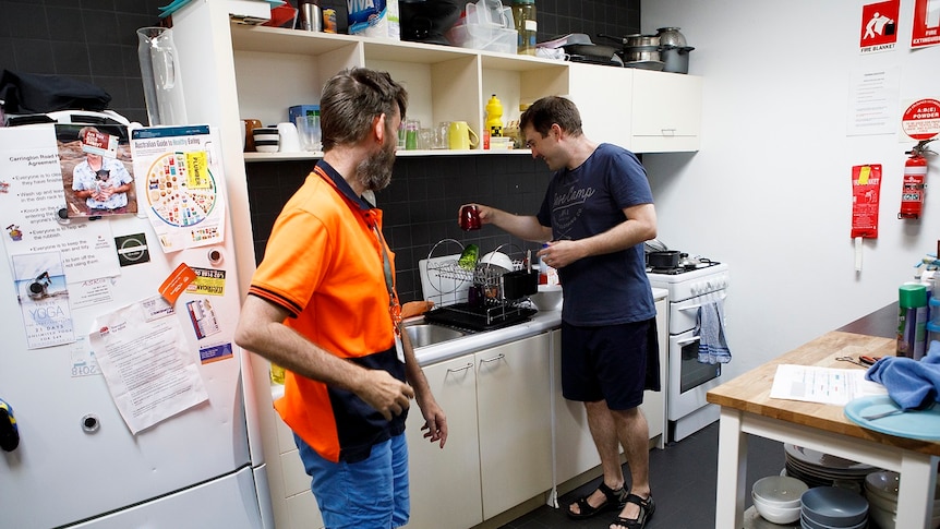 A man puts away cups from a kitchen drainer while another man watches for a story on share housing.