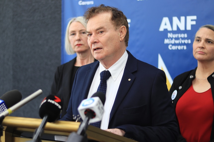 A man in a suit speaks at a press conference watched on by two women. 