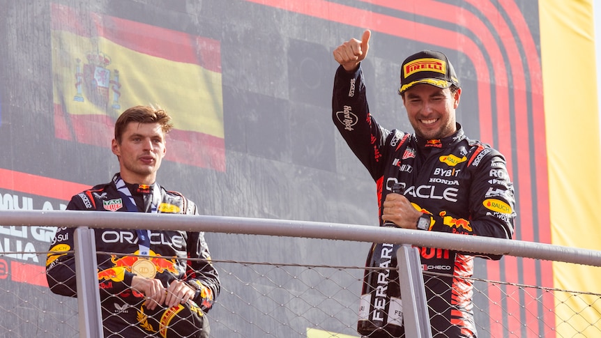 Two F1 drivers, one giving a thumbs up, stand on a podium, smiling and celebrating.