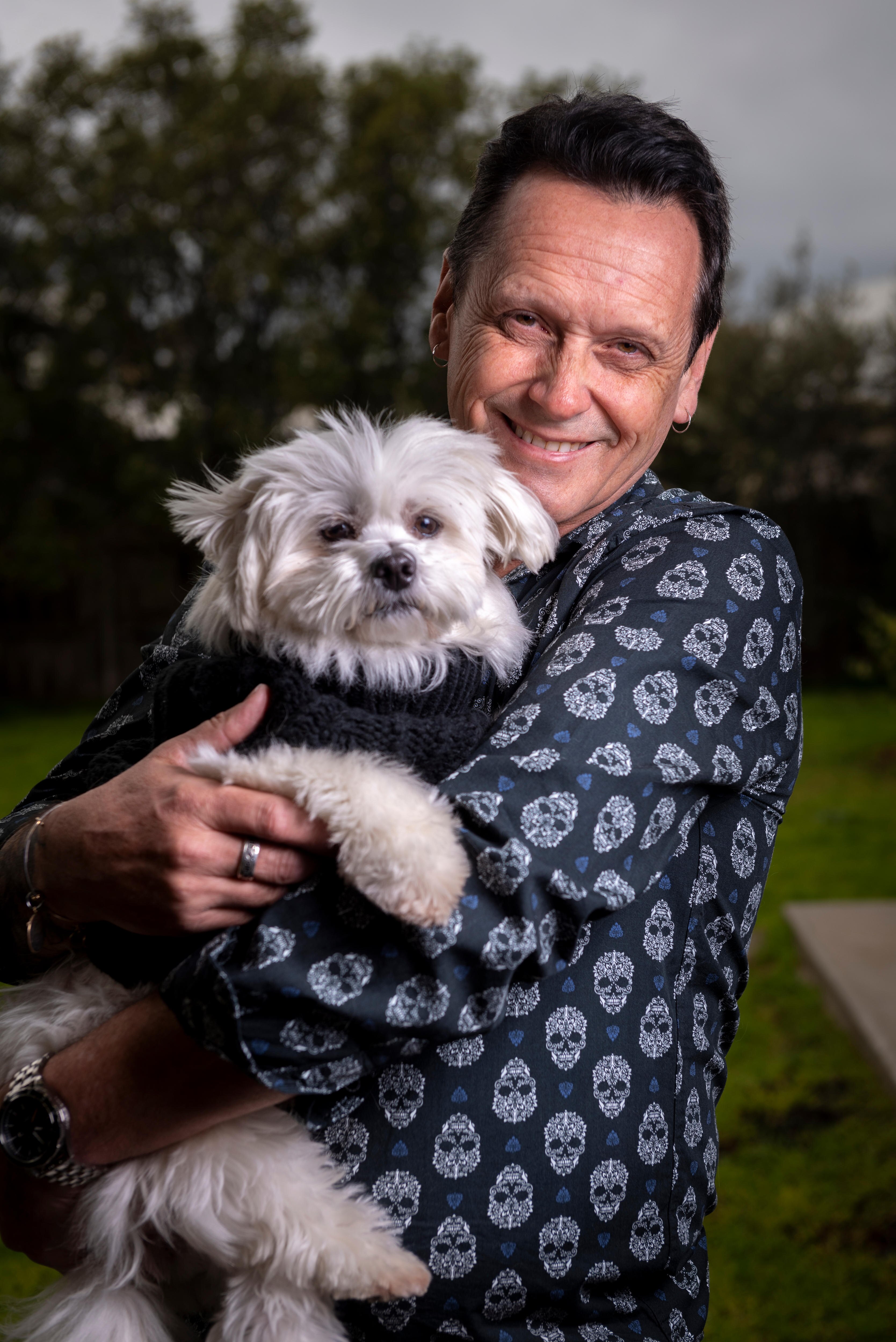 Andy Meddick smiles, dressed in a dark blue shirt as he holds a small dog dressed in a warm coat, under gray skies.