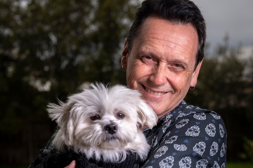 Andy Meddick smiles, dressed in a dark blue shirt as he holds a small dog dressed in a warm coat, under gray skies.