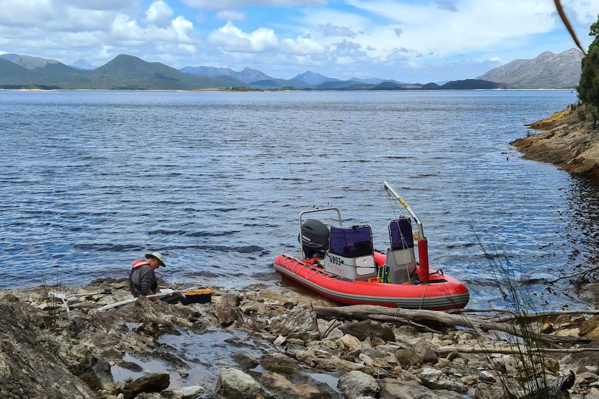 A large inland lake. A man sits on a rocky shoreline where a small boat is moored.