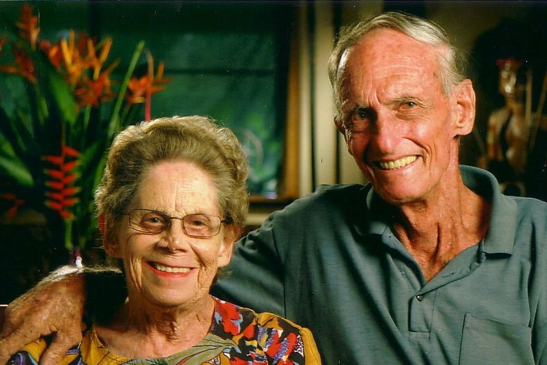 A older woman and a man sit smiling together in front of flowers in the background.