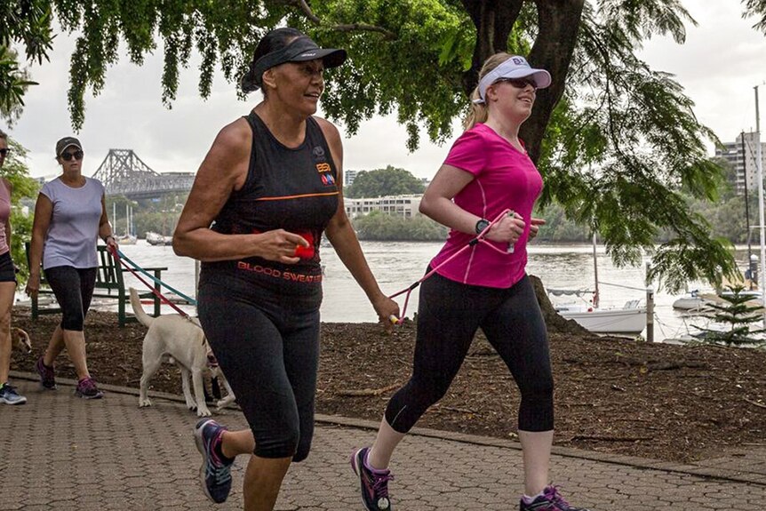 Two women running together in a park, holding a pink tether between them.