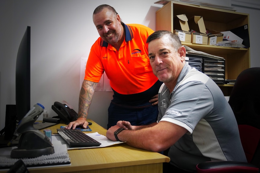 A man in a polo shirt sits at a desk near another man who is standing and smiling.