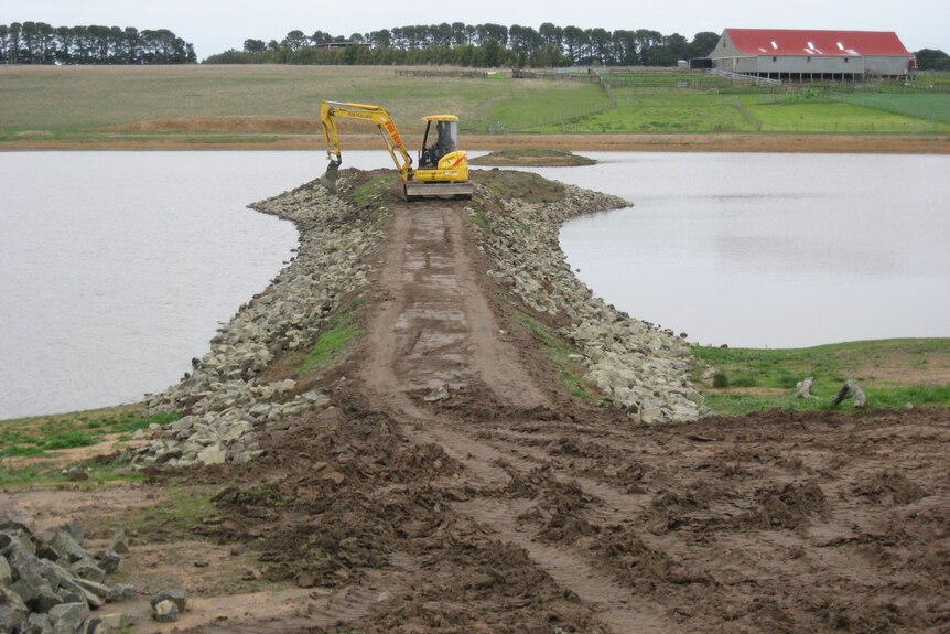 an excavator moving soil around on a dry land bridge between two bodies of water. house in the background.