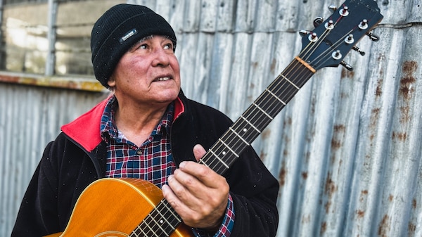 A man wearing a black jacket, plaid shirt and black beanie holds an acoustic guitar.