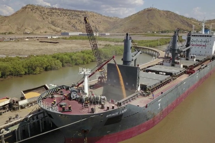 An industrial ships storage hold is filled with grain from a crane mechanism, while docked at a port.