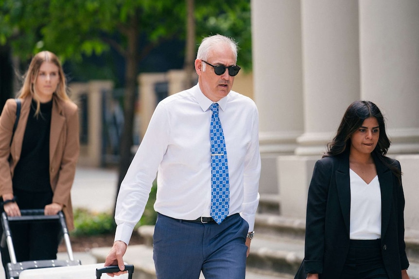 Francis Wark's lawyer Darryl Ryan walking outside court wearing sunglasses and a blue tie, with women to his left and right.
