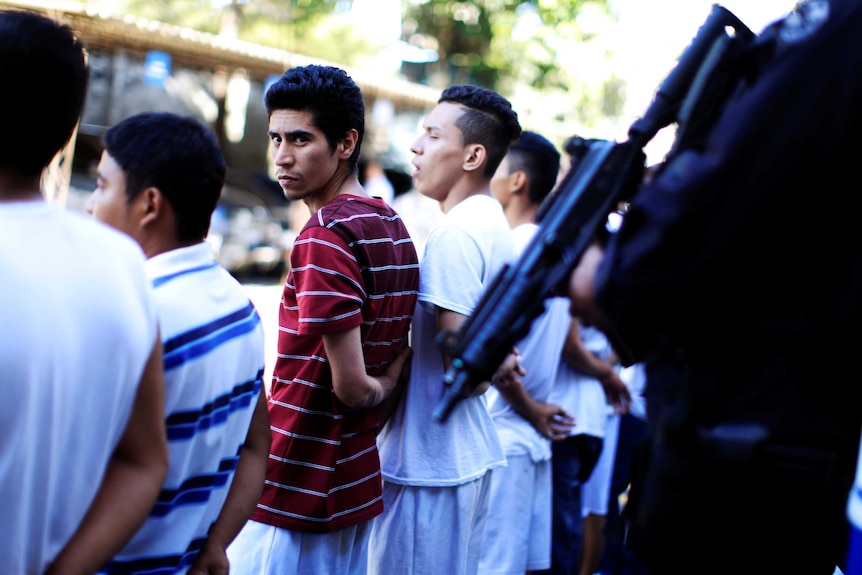 Gang members line up with hands behind their back as a policeman holding a rifle stands guard