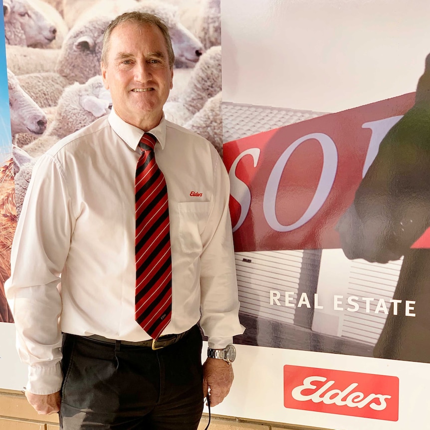 A man is standing up in a suit with a red tie next to a sold sign