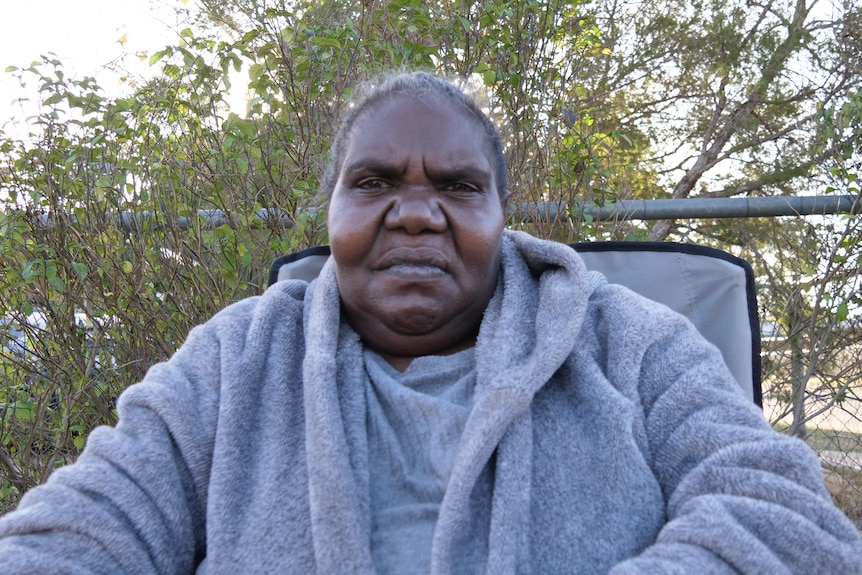An Aboriginal woman in a grey jacket sits on a chair in front of a shrub