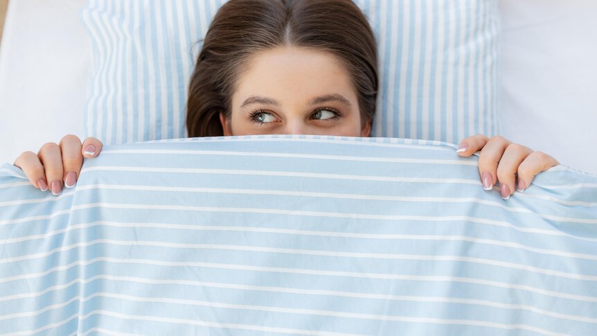 A woman lies in bed with striped blue covers pulled up to her eyes. She looks to the side with a playful expression.