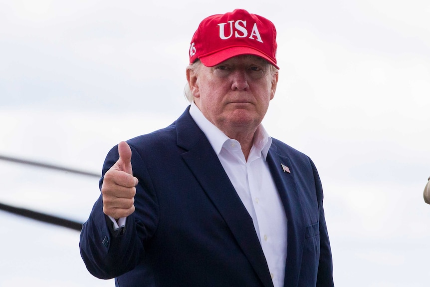 Donald Trump gives a thumbs up while wearing a red baseball cap saying "USA".