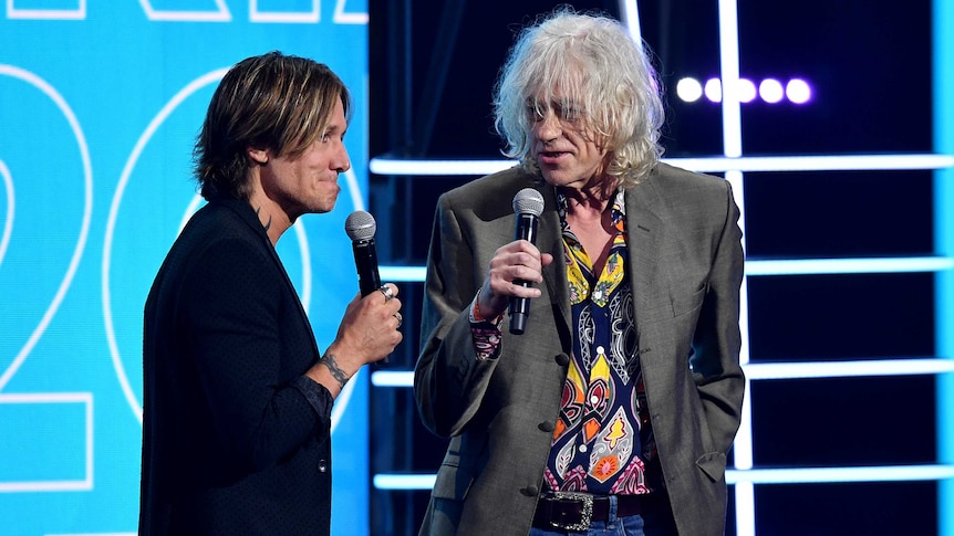Keith Urban wears an awkward expression on his face while Bob Geldof speaks into a microphone on stage.