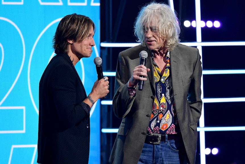Keith Urban wears an awkward expression on his face while Bob Geldof speaks into a microphone on stage.