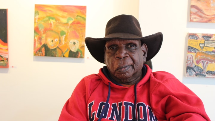 An older Aboriginal man in a cowboy hat sits in a wheelchair, bright paintings behind him. He's wearing a red London jumper