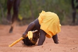 A baby boy in traditional Aboriginal paint and dress leans over with his head on the ground