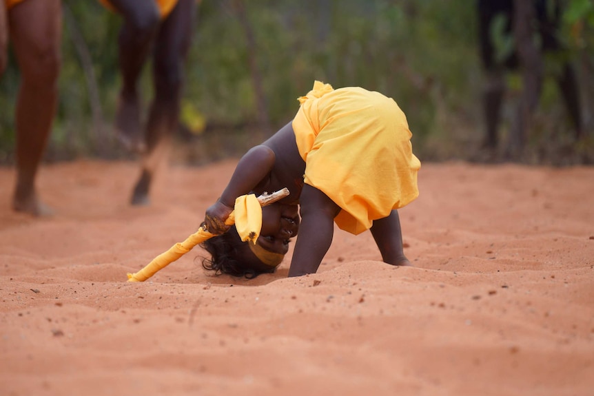A baby boy in traditional Aboriginal paint and dress leans over with his head on the ground.