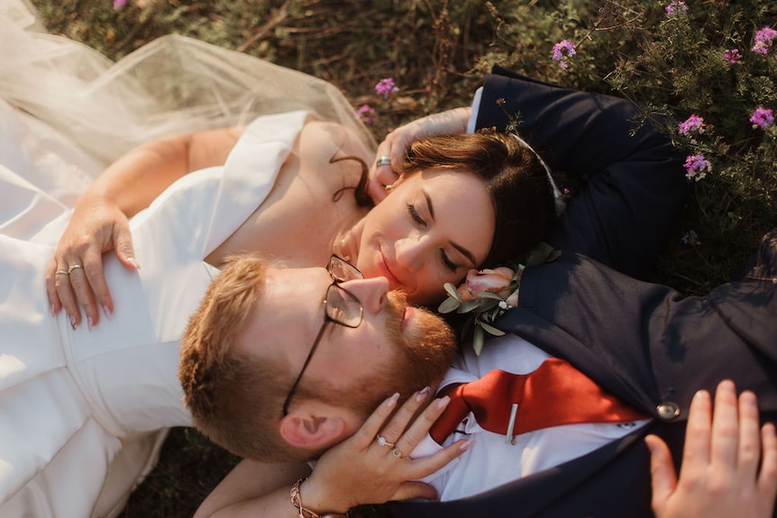 A bride and her groom lie on grass, sharing a loving embrace.