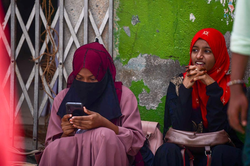 Two young girls in Kerala sitting by a wall, one of them looking at a phone